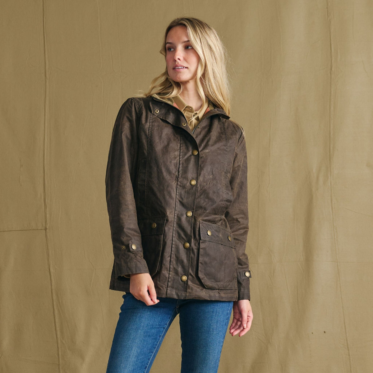 Woman wears the Tom Beckbe Women’s Blakeley Jacket with blue jeans, posing against a tan background.