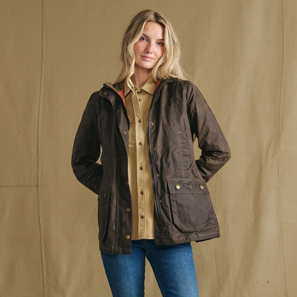 Woman in Tom Beckbe’s Blakeley Jacket stands before a beige backdrop, styled with a tan shirt and blue jeans.