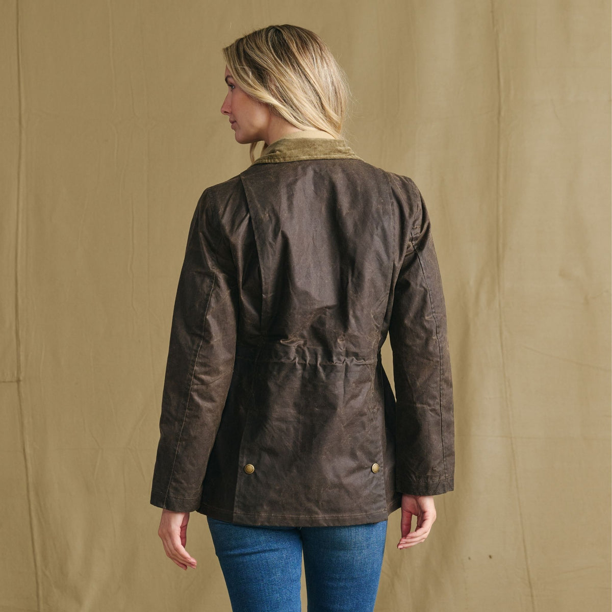 Woman in Tom Beckbe Women’s Blakeley Jacket and jeans, back to camera, against beige background.