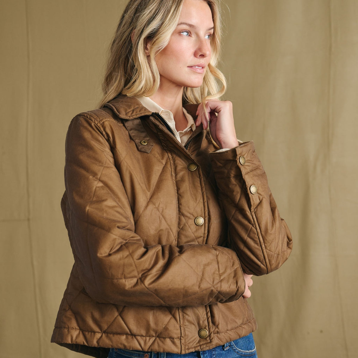 Woman wearing a brown quilted jacket stands in front of a beige background, looking to the side.