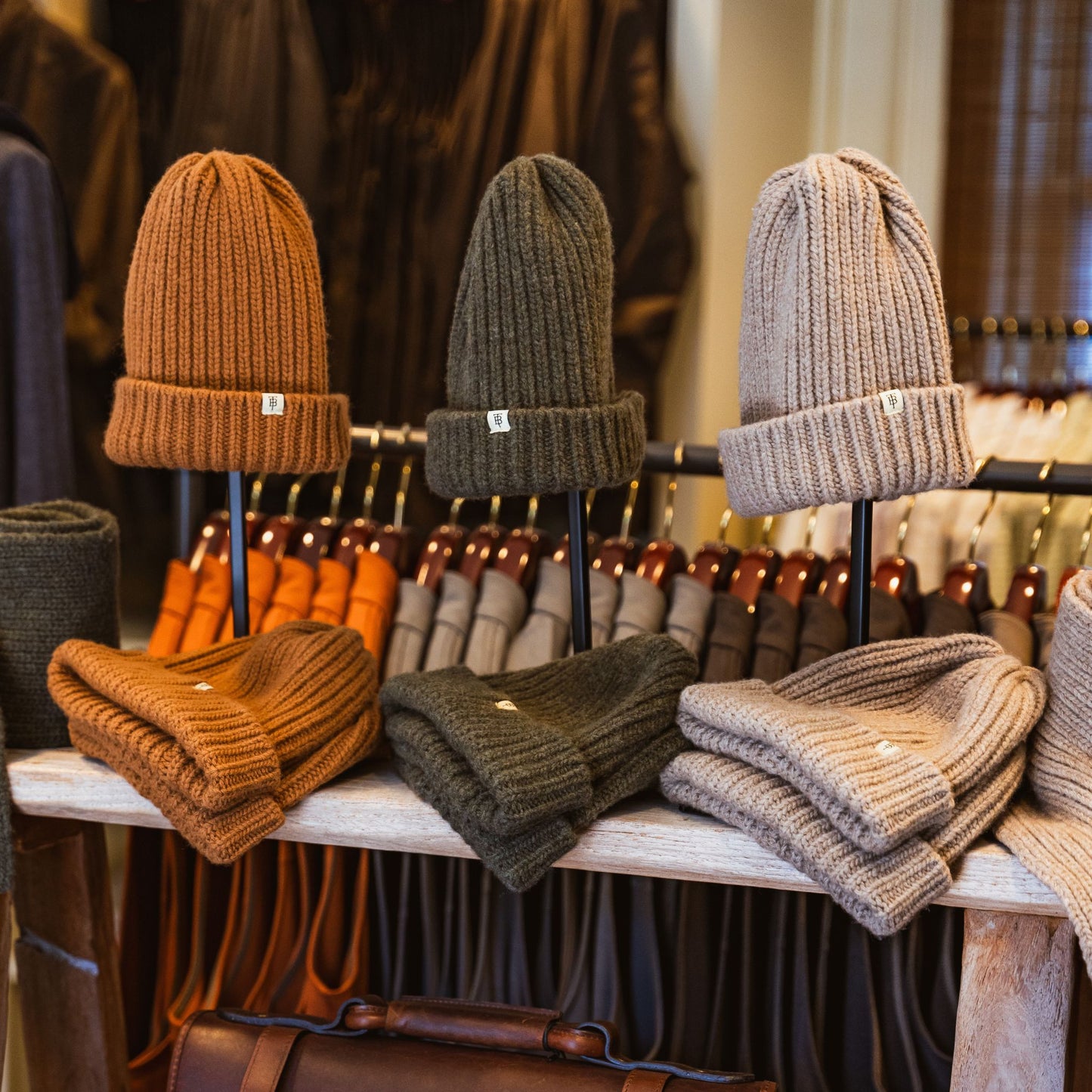 Three Tom Beckbe Wool Caps in brown, green, and beige are displayed on a wooden table in-store.