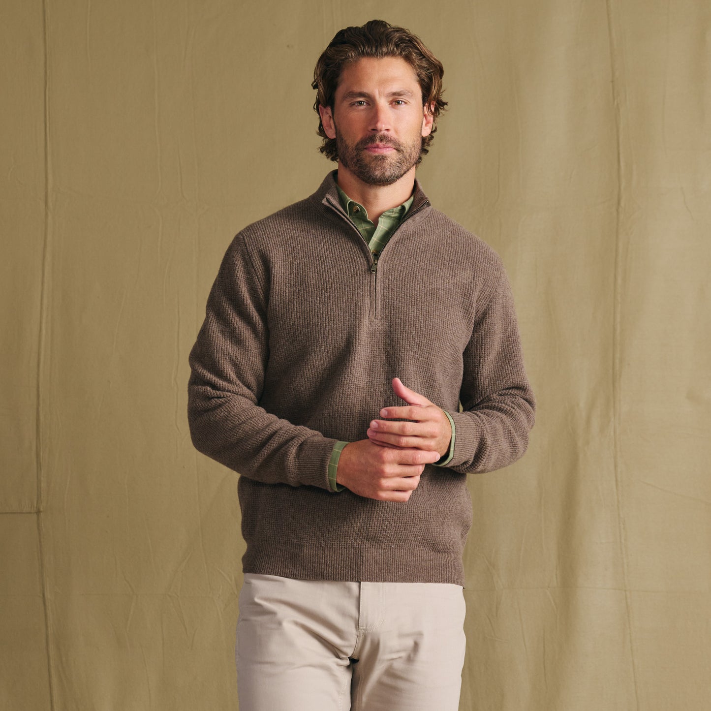 Man wears Tom Beckbe Farris Waffle Knit Quarter Zip over green shirt, standing before a beige fabric backdrop.