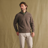 Man wears Tom Beckbe Farris Waffle Knit Quarter Zip over a green shirt, beige pants, and stands by a tan fabric backdrop.