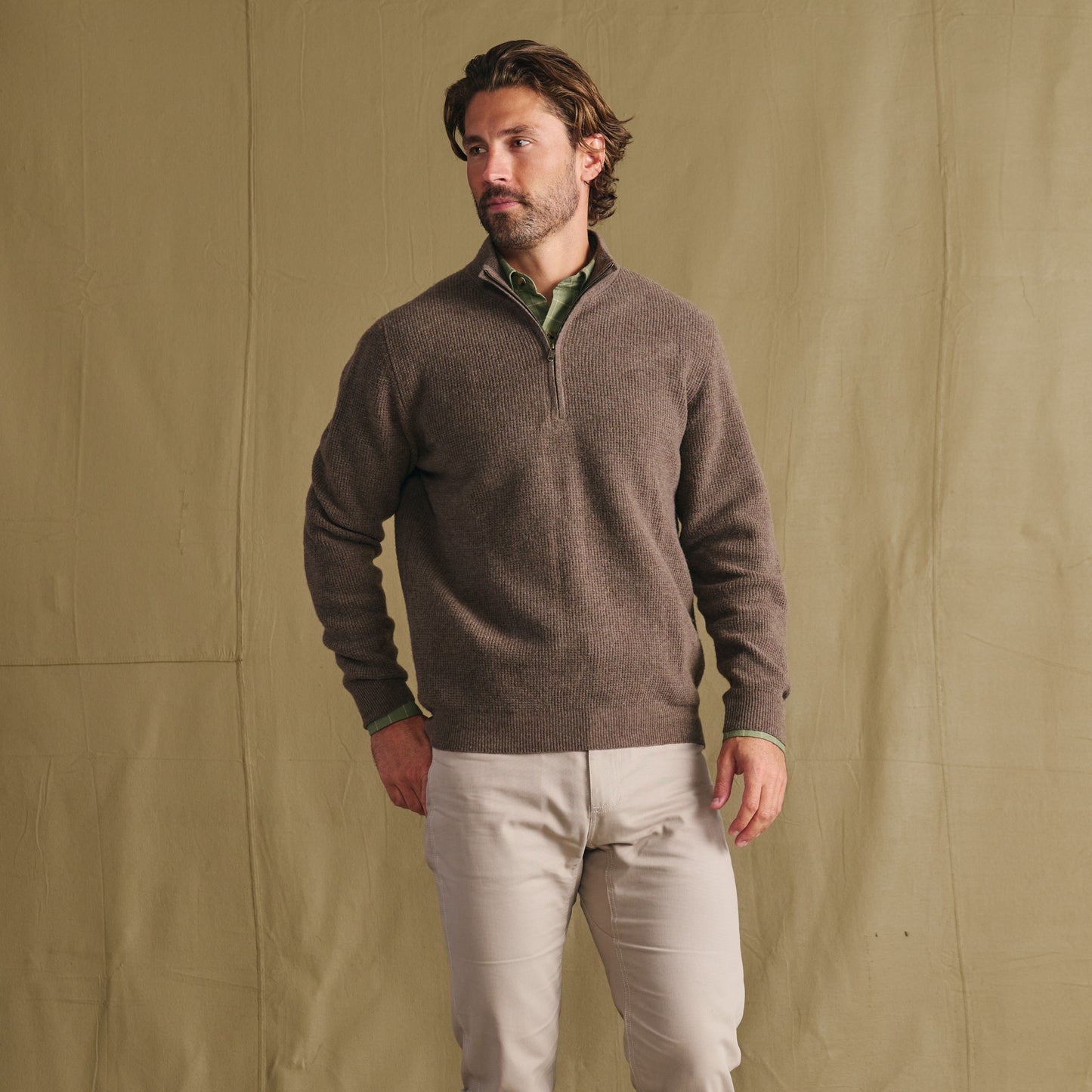 Man wears Tom Beckbe Farris Waffle Knit Quarter Zip over a green shirt, beige pants, and stands by a tan fabric backdrop.