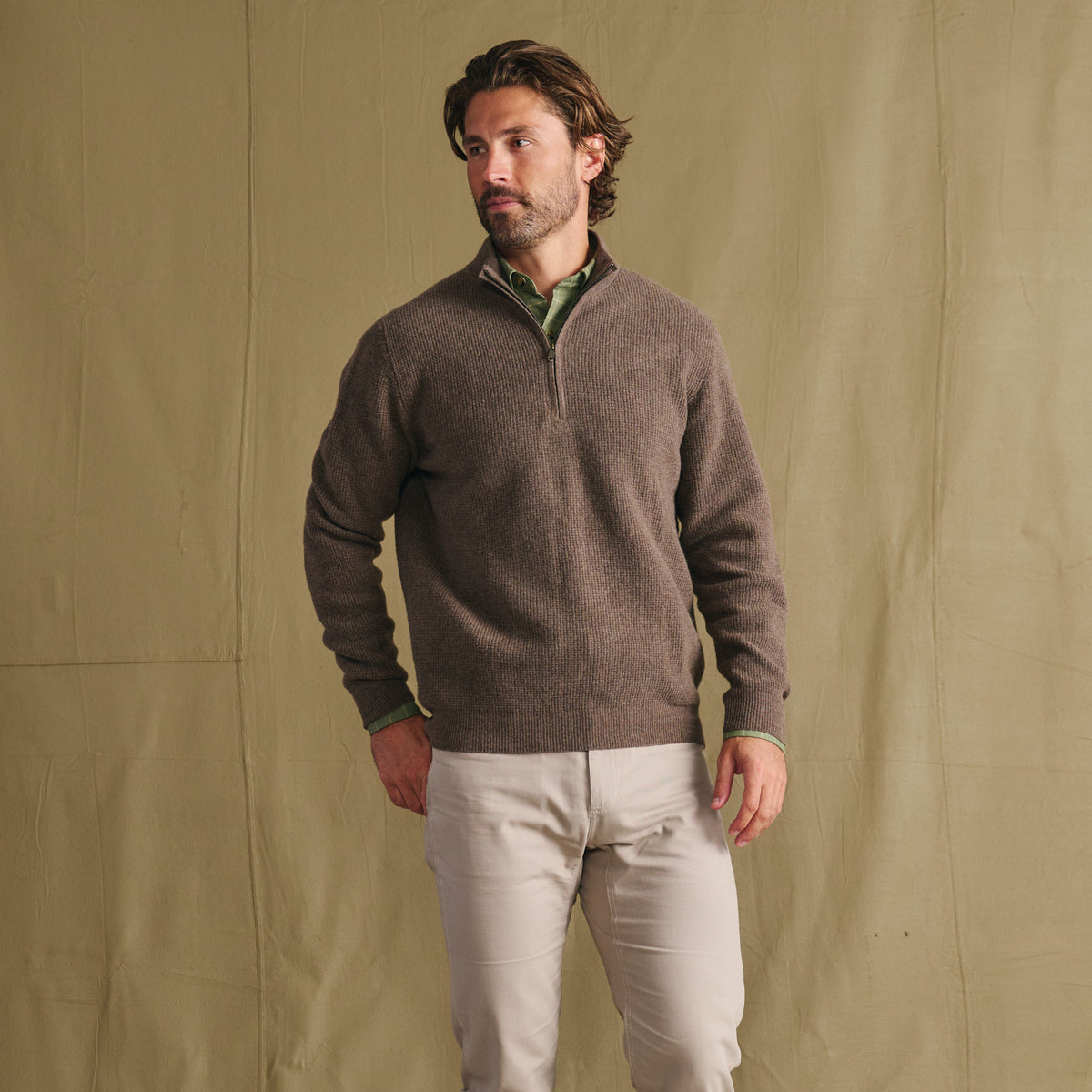 Man wears Tom Beckbe Farris Waffle Knit Quarter Zip over a green shirt, beige pants, and stands by a tan fabric backdrop.