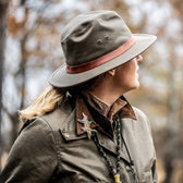 Person wearing the Tom Beckbe DU Field Hat and an olive green outdoor jacket, looking to the side in a wooded area.
