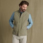 Man wearing a light green quilted vest over a blue button-down shirt and beige pants, standing with hands in pockets.