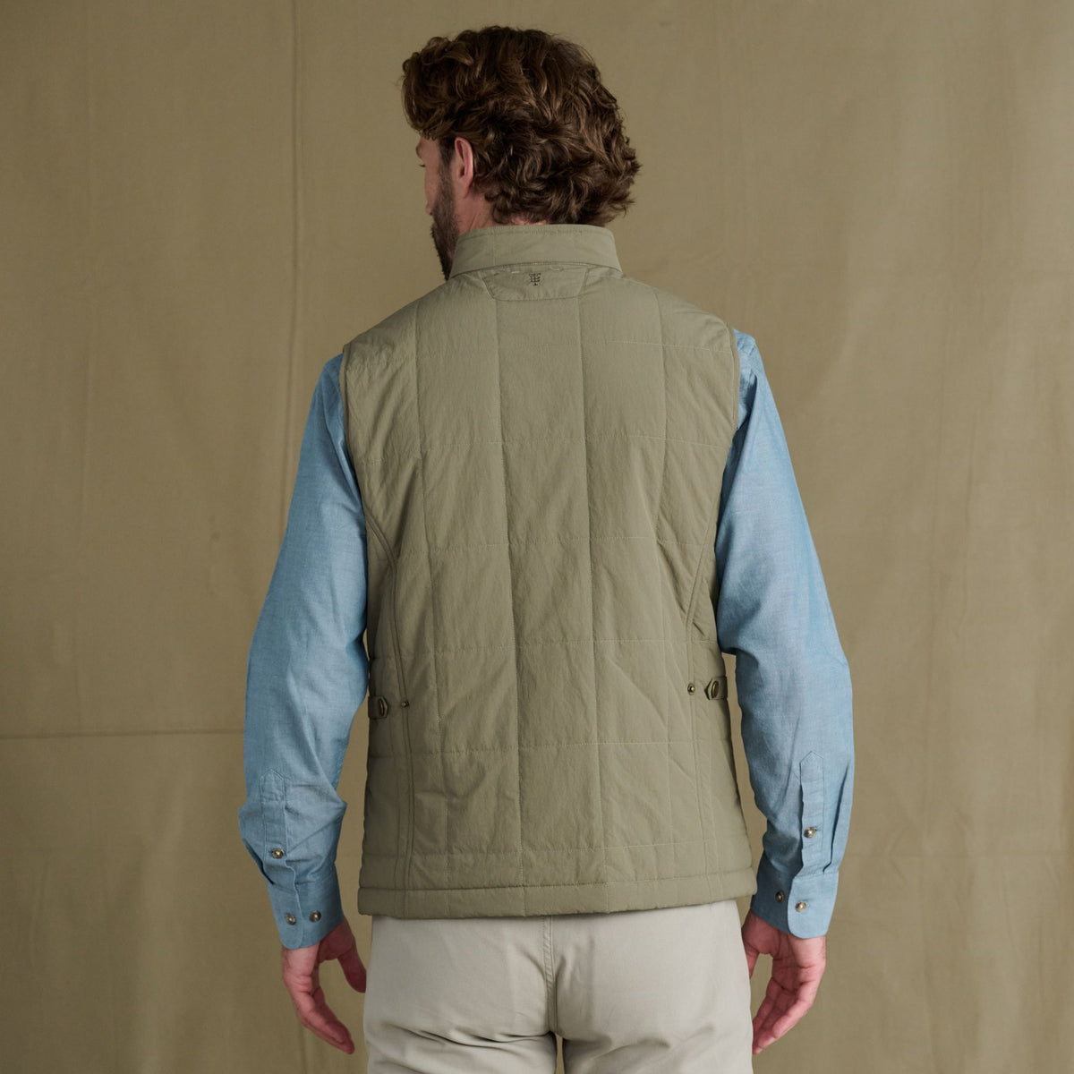 Man facing away, wearing a light green quilted vest over a blue shirt and beige pants, standing against a tan background.