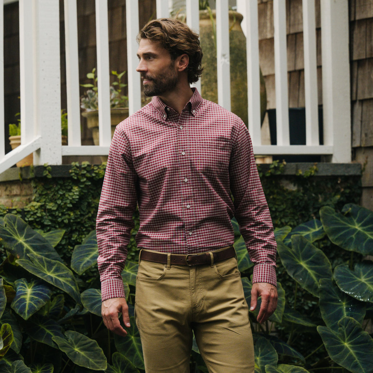 Man wearing a red checkered shirt and khaki pants stands outdoors in front of lush green plants and a white railing.