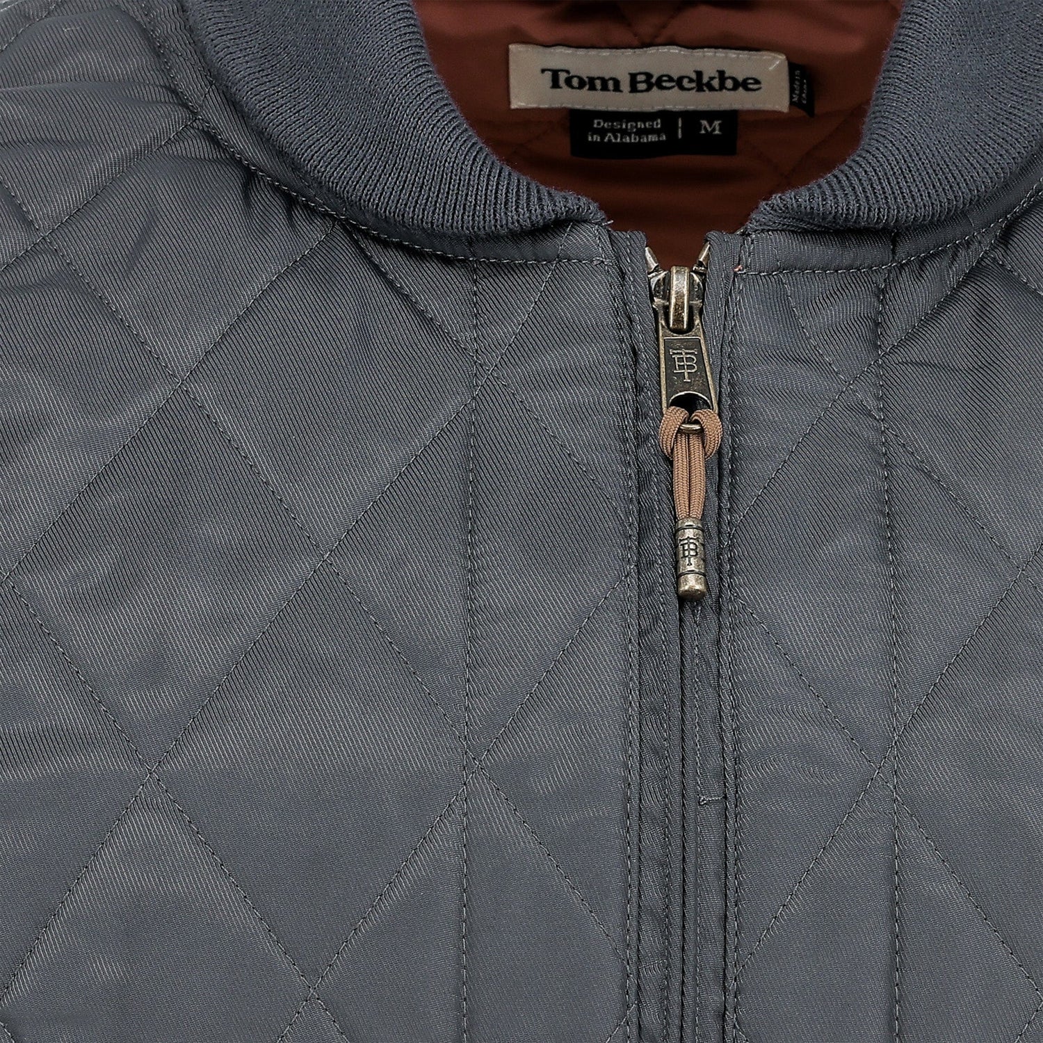 Close-up of a gray quilted jacket with a front zipper and a visible Tom Beckbe brand label inside the collar.