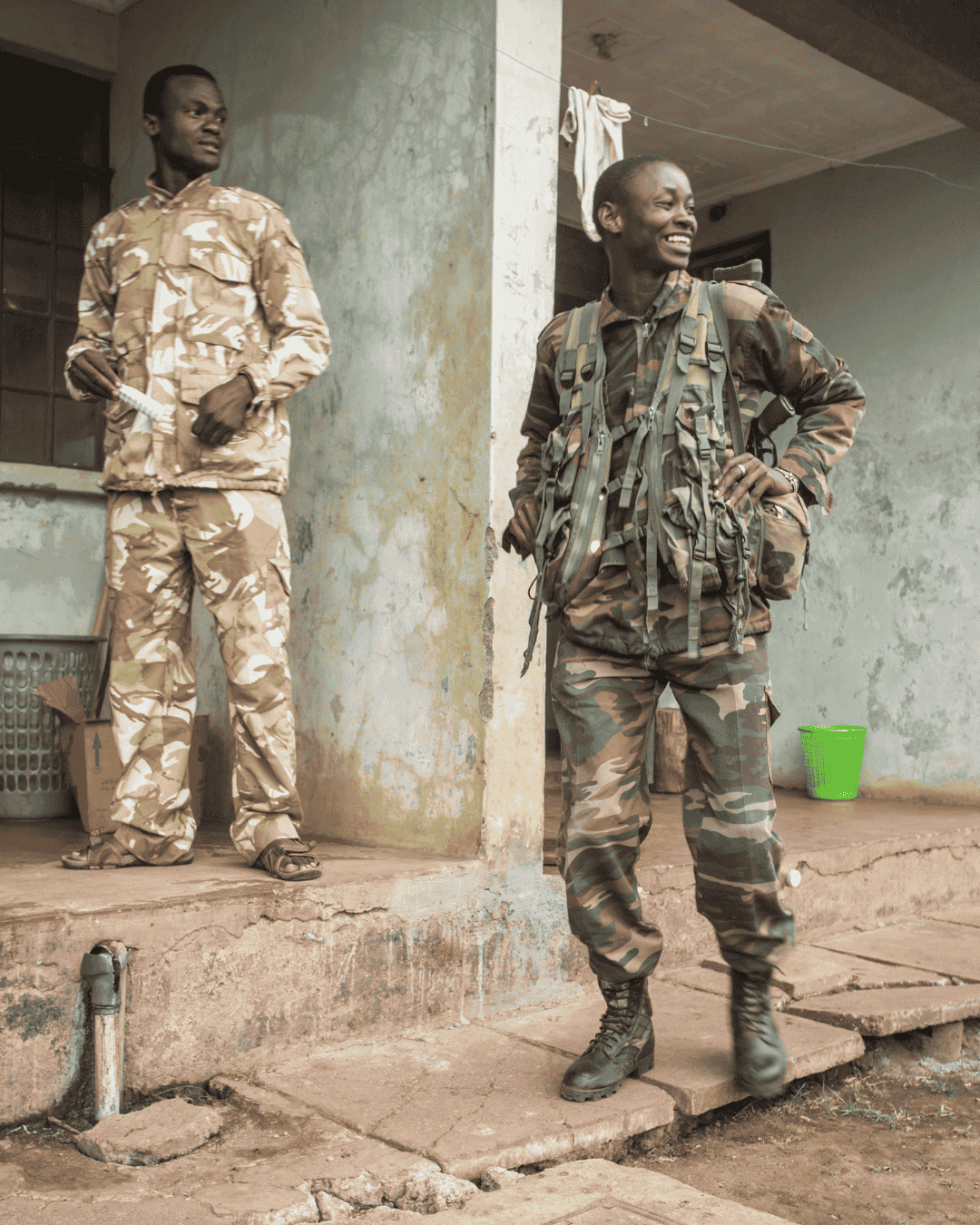 Two people stand on a porch wearing camouflage uniforms. One person is smiling and looking to the side.