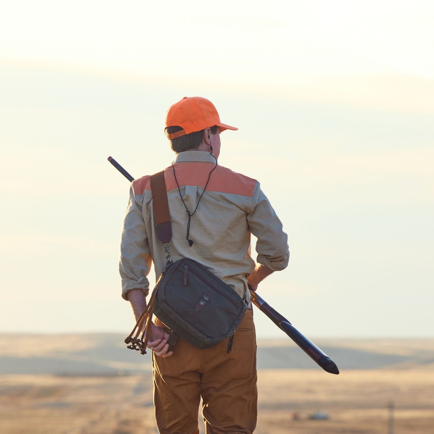 A person in a Tom Beckbe Uplander Shirt and orange cap carries a rifle across a vast landscape at sunset.