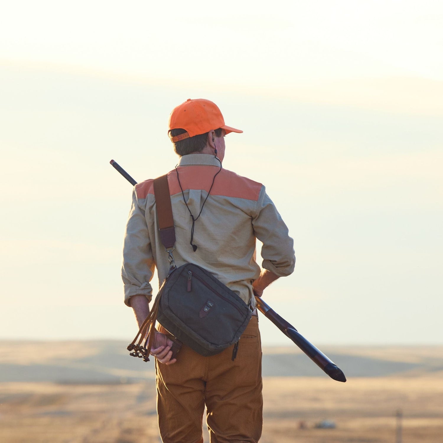 A person in a Tom Beckbe Uplander Shirt and orange cap carries a rifle across a vast landscape at sunset.