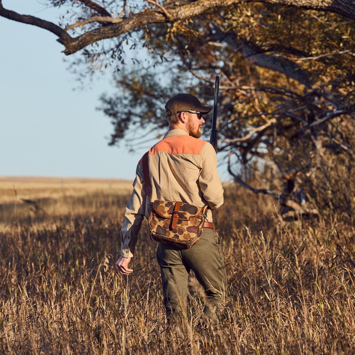 A person in a Tom Beckbe Uplander Shirt walks with a rifle through a grassy field with trees in the background.