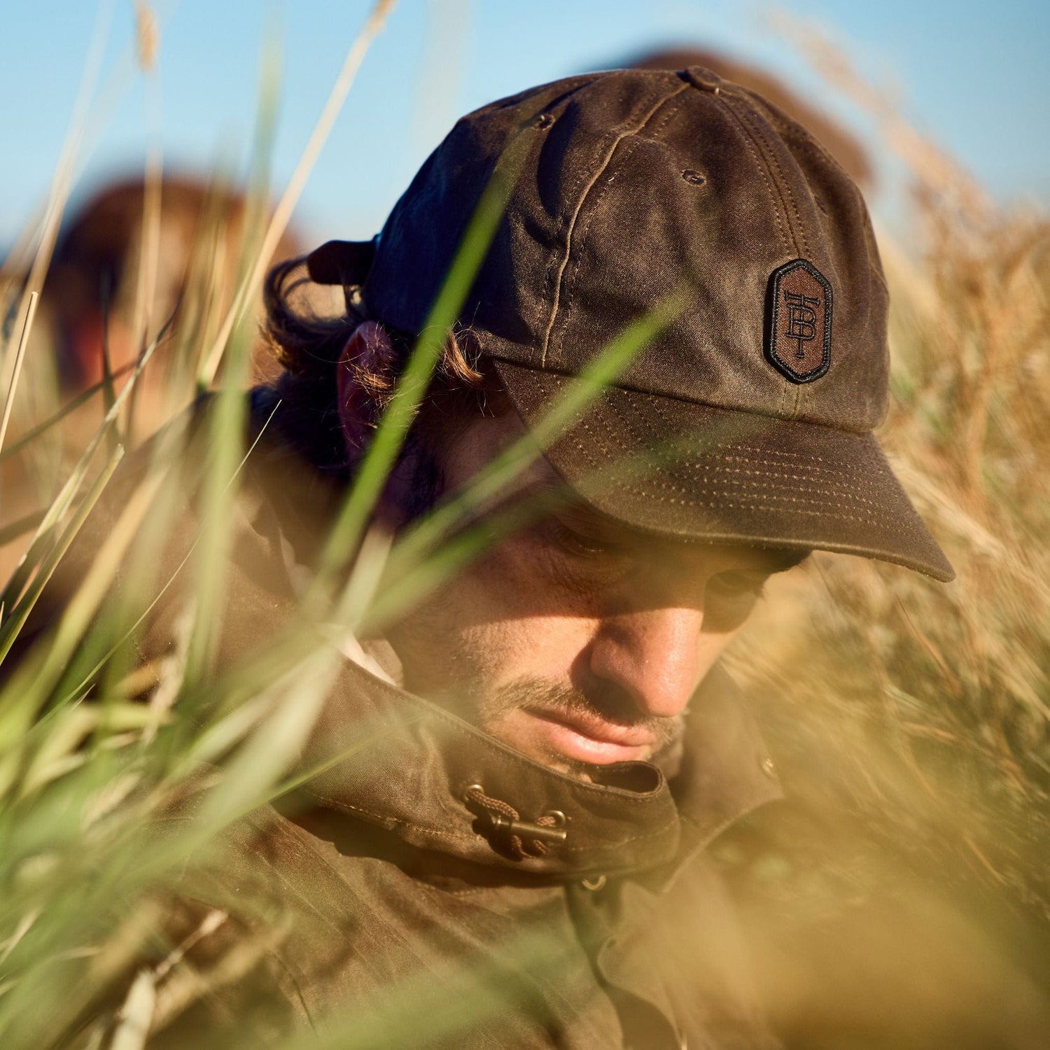 A person in a Tom Beckbe Waxed Cotton 6-Panel Baseball Hat and jacket stands in tall grass, with another person behind.