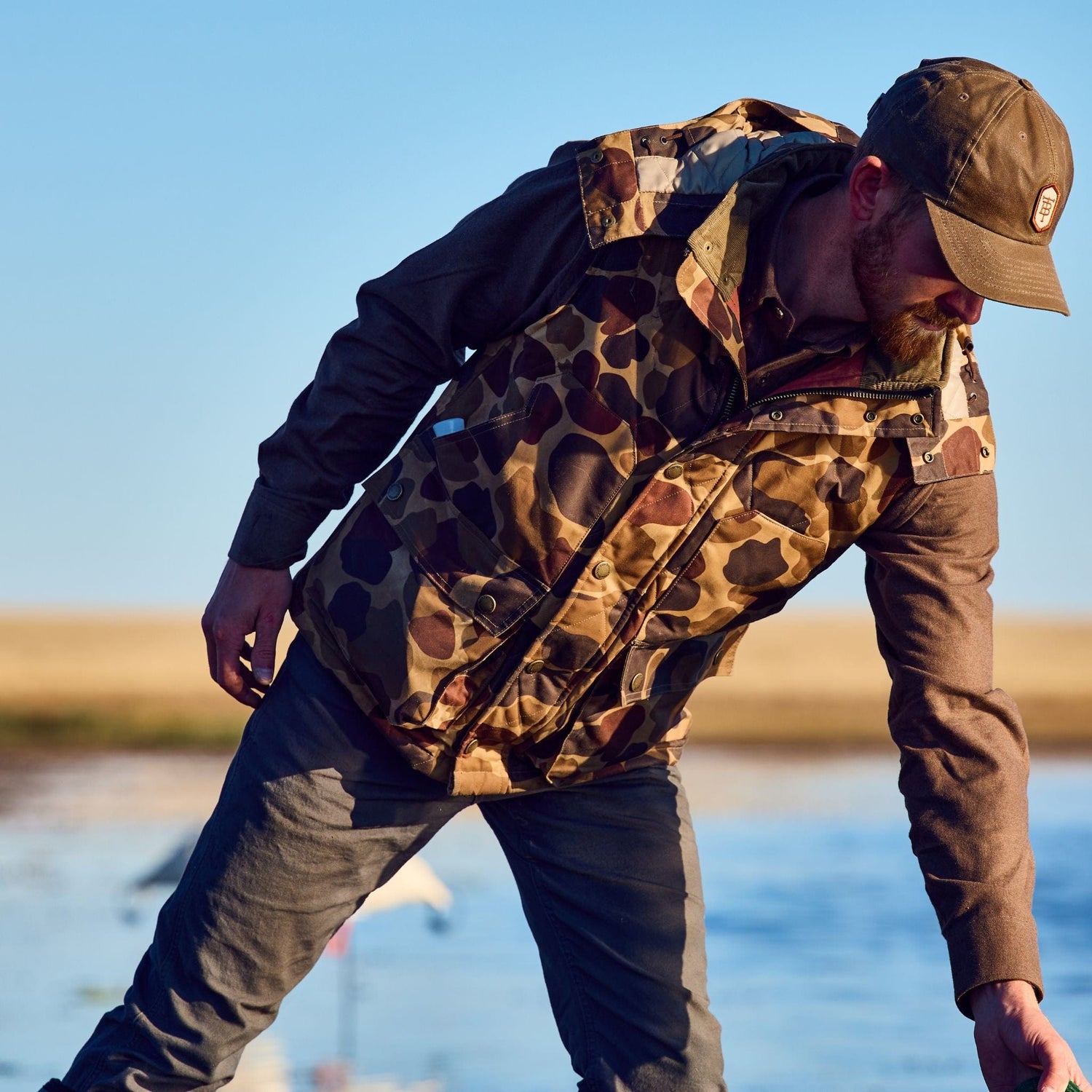 A person in a camouflage jacket and Tom Beckbe hat kneels by water with grass behind.
