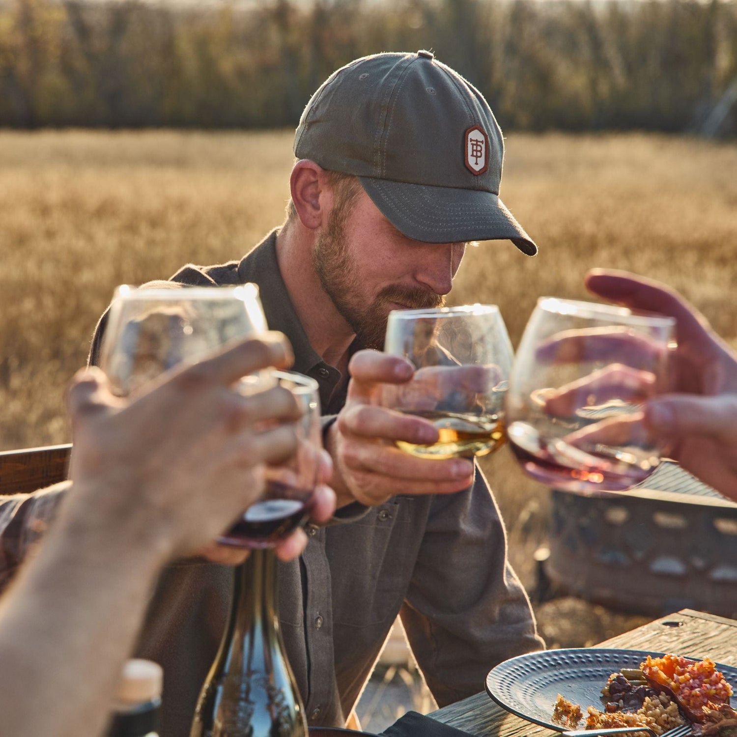 People clinking glasses outdoors over a meal with one wearing a Tom Beckbe Waxed Cotton 6-Panel Baseball Hat, fields behind.