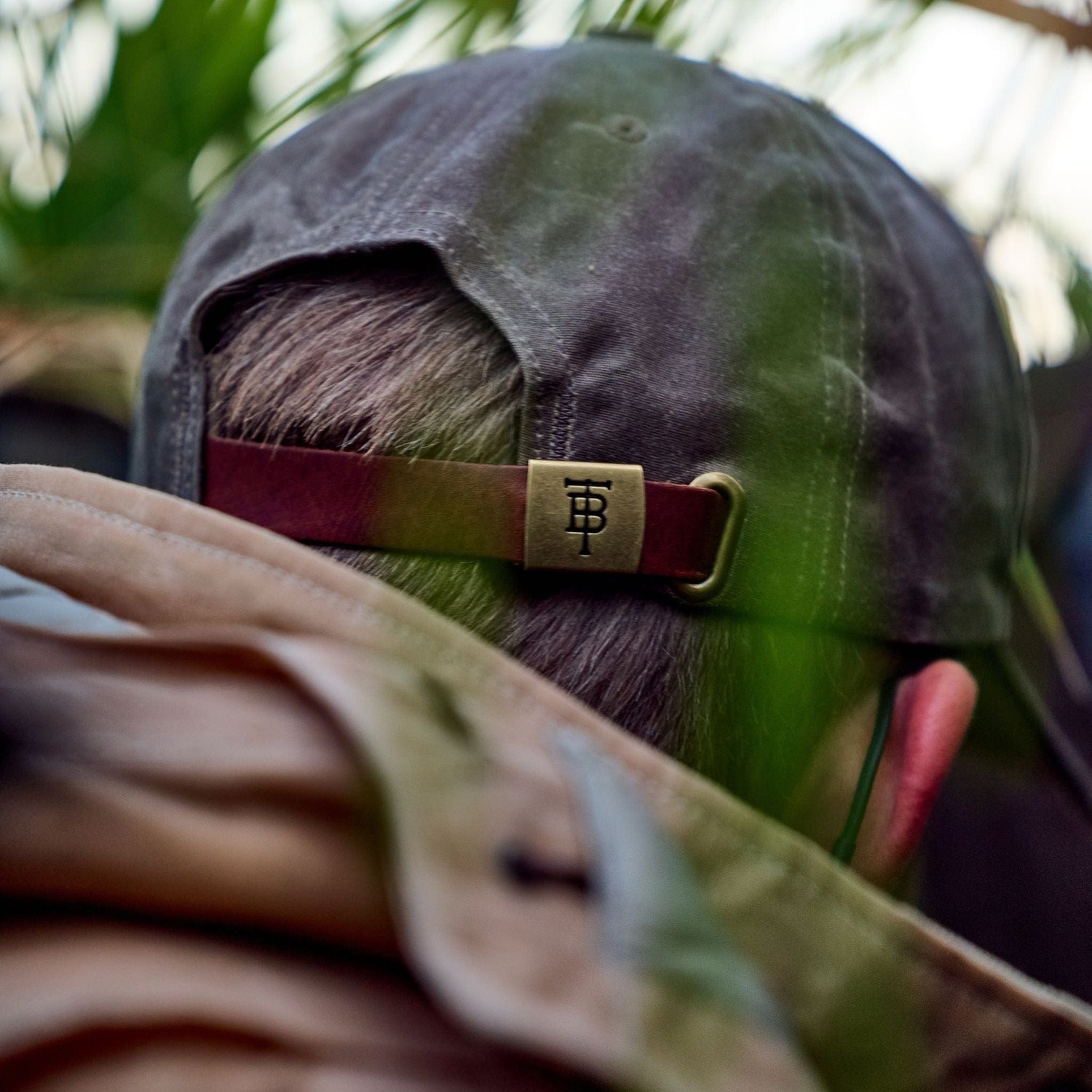 Person in a Tom Beckbe Waxed Cotton 6-Panel Baseball Hat amidst foliage, looking away from the camera.