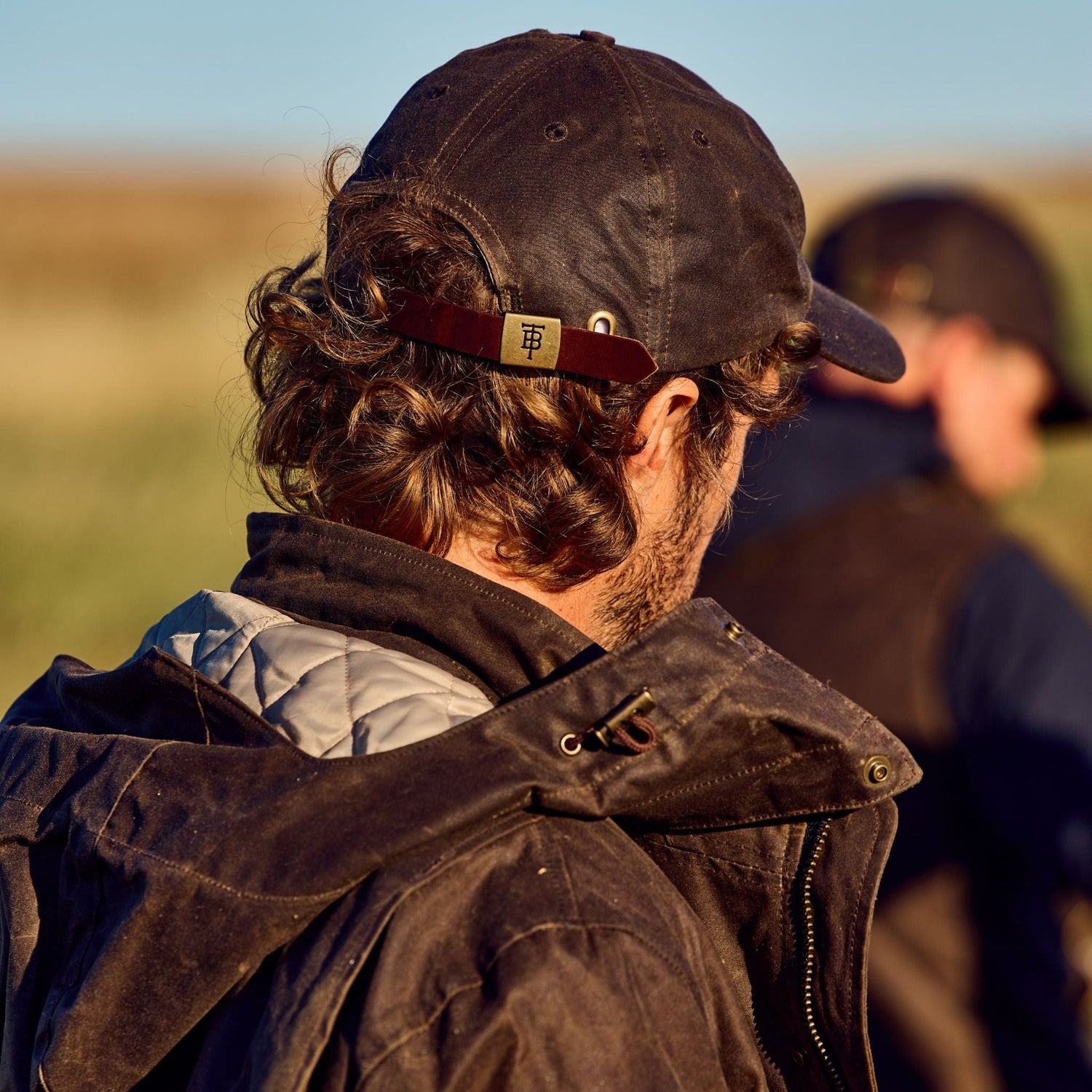 Two people in outdoor jackets walk in a field wearing Tom Beckbes Waxed Cotton 6-Panel Baseball Hat under a clear sky.