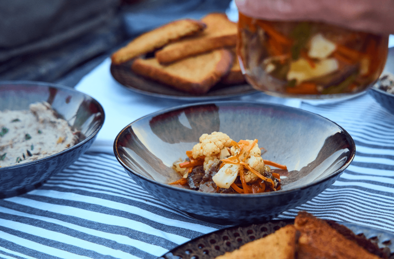 A bowl of cauliflower, carrots, and meat on a striped tablecloth with toast in the background.