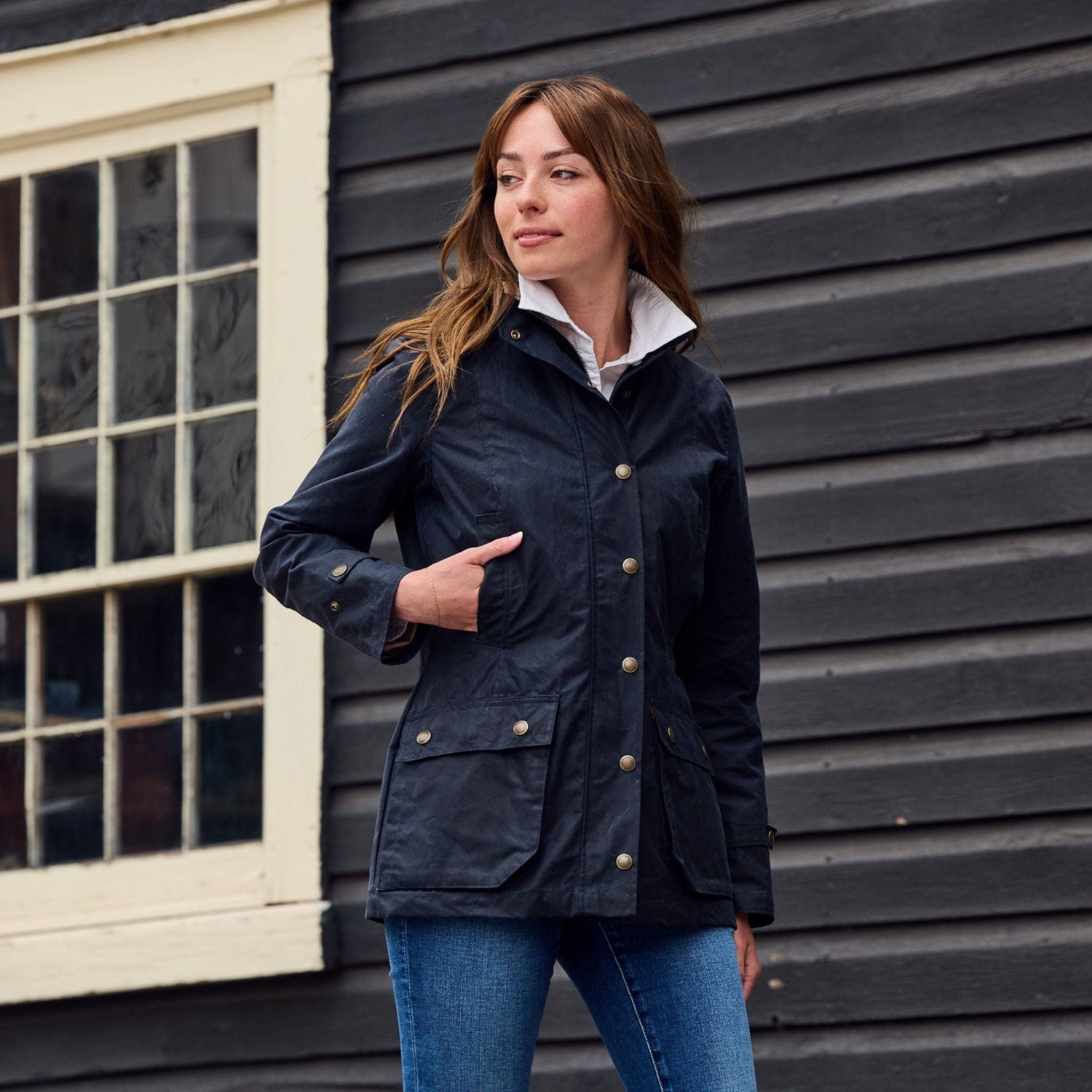 A woman in a Tom Beckbe Womens Blakeley Jacket and blue jeans stands outside a black building with white-trimmed windows.