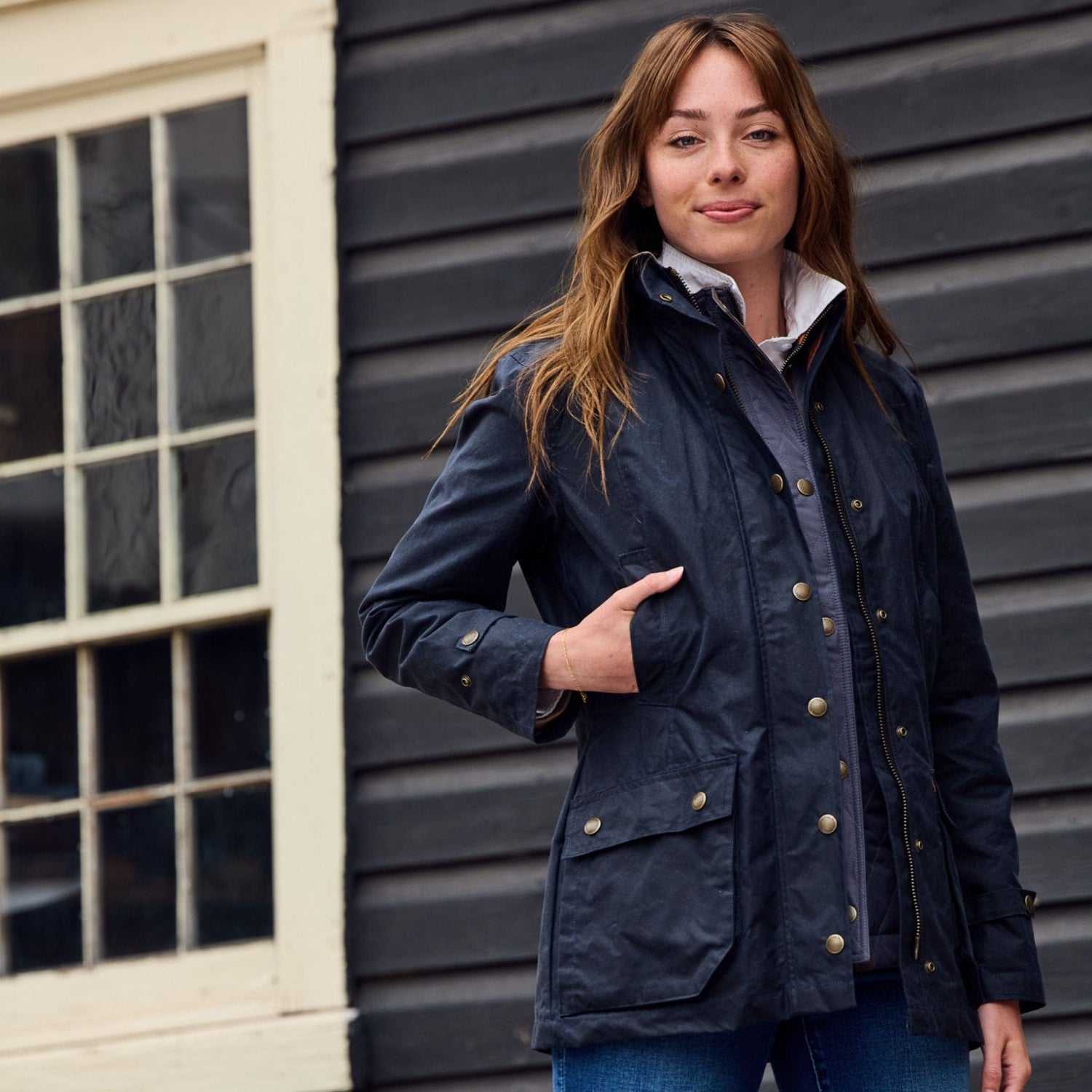 A woman in a Tom Beckbe Womens Blakeley Jacket stands by a window and dark wooden wall, her hands in the pockets.