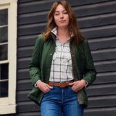 A woman stands confidently in a Tom Beckbe green Womens Suffolk Sport Shirt and jeans before a dark wooden wall with a window.