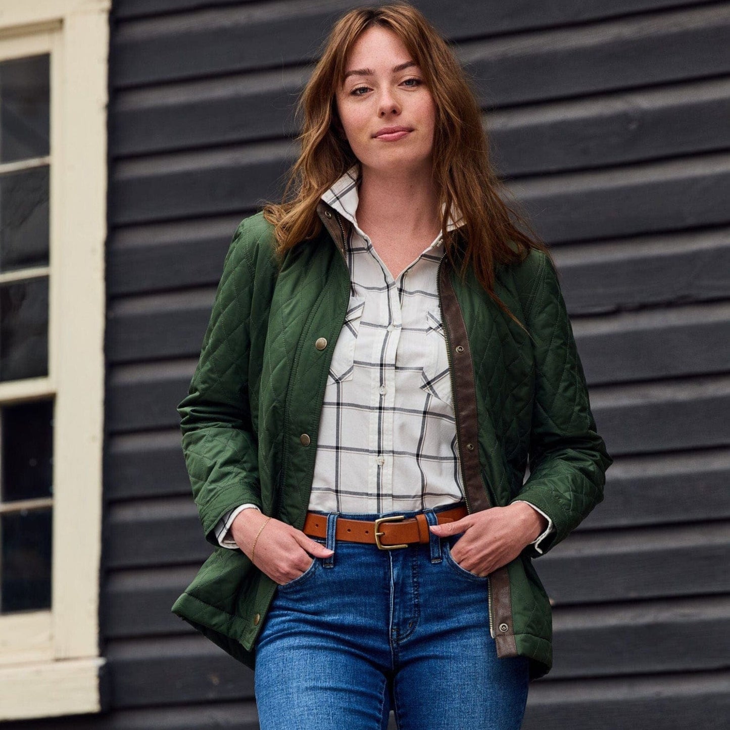 A woman stands confidently in a Tom Beckbe green Womens Suffolk Sport Shirt and jeans before a dark wooden wall with a window.