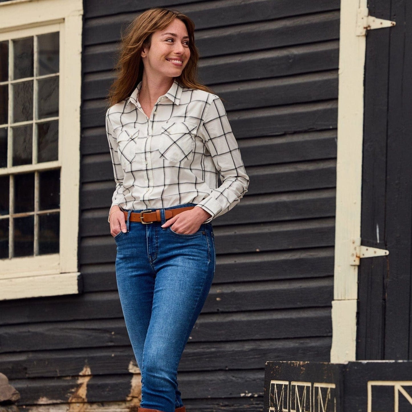 A person in a Womens Suffolk Sport Shirt by Tom Beckbe stands before a dark wooden building, smiling and looking away.