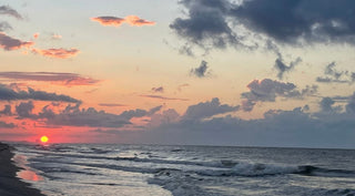 Sunset over a beach with waves rolling onto the shore. The sky is filled with clouds and the sun is near the horizon.