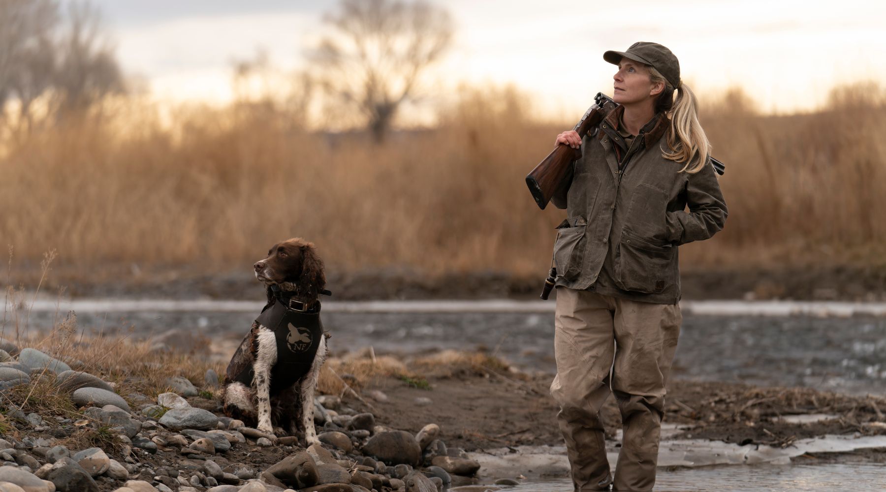 A woman carrying a rifle walks near a river with a dog beside her; both are dressed in outdoor gear.