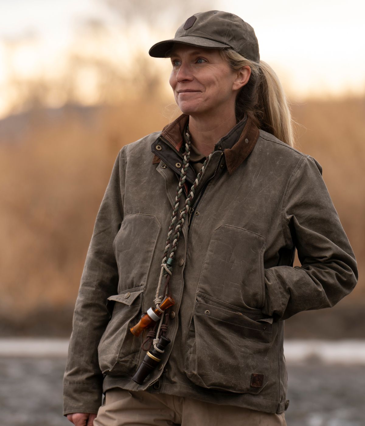 Woman in a green jacket and cap stands outdoors with a whistle and lanyard around her neck, hands in her pockets.