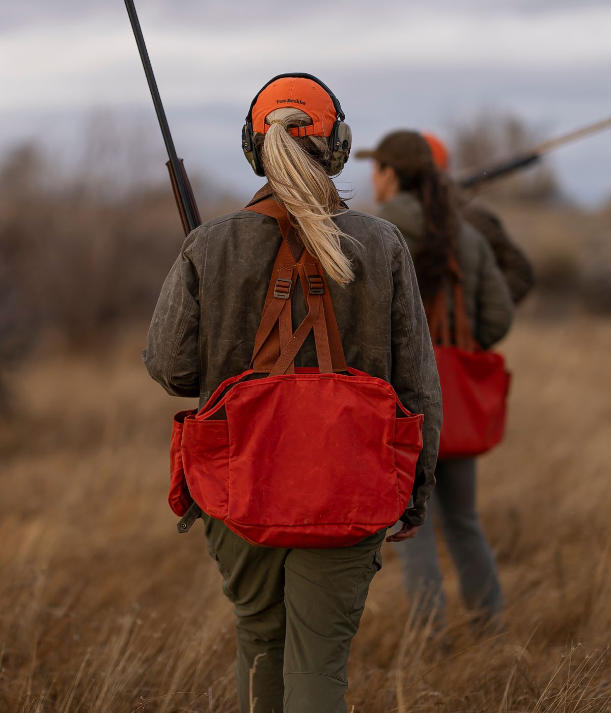 Two people walk through tall grass carrying guns, wearing orange hats and large red bags on their backs.