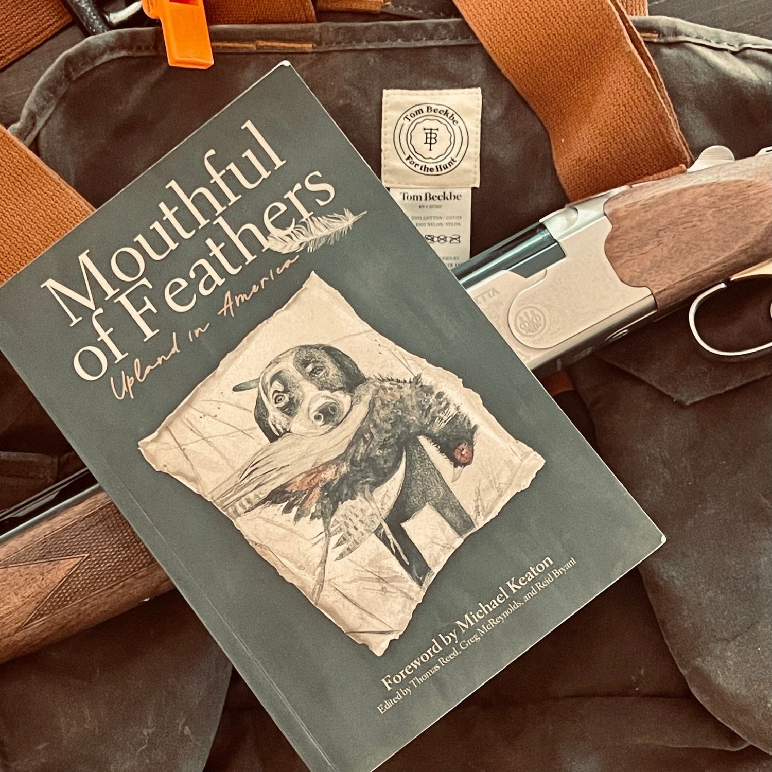 A book titled Mouthful of Feathers rests on a hunting bag with a shotgun, shells, and a bottle of whiskey.