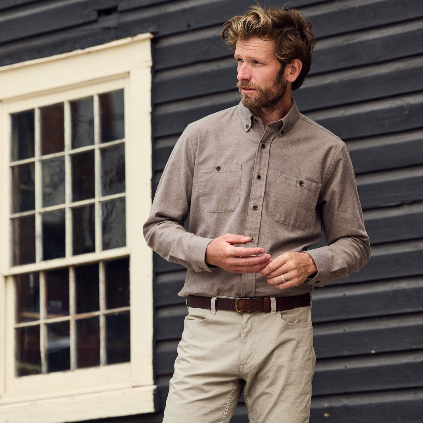 A man in a Tom Beckbe Dixon Brushed Cotton Twill Shirt and beige pants stands outside by a black building with white windows.