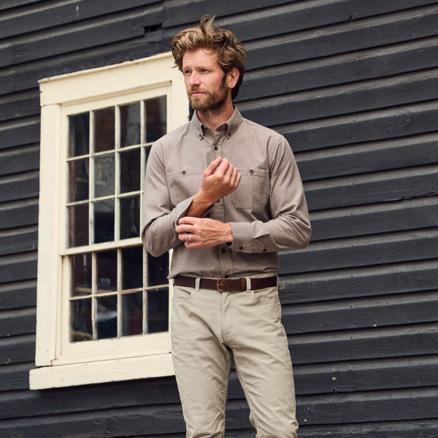 A man in a Tom Beckbe Dixon Brushed Cotton Twill Shirt adjusts his cuff by a white-trimmed window.