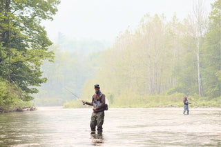 Two people stand in a shallow river fly fishing, surrounded by trees and greenery on a hazy day.