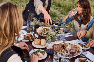 People dining outdoors at a table with wine, water, bread, and various dishes. A person pours wine while others eat and drink.