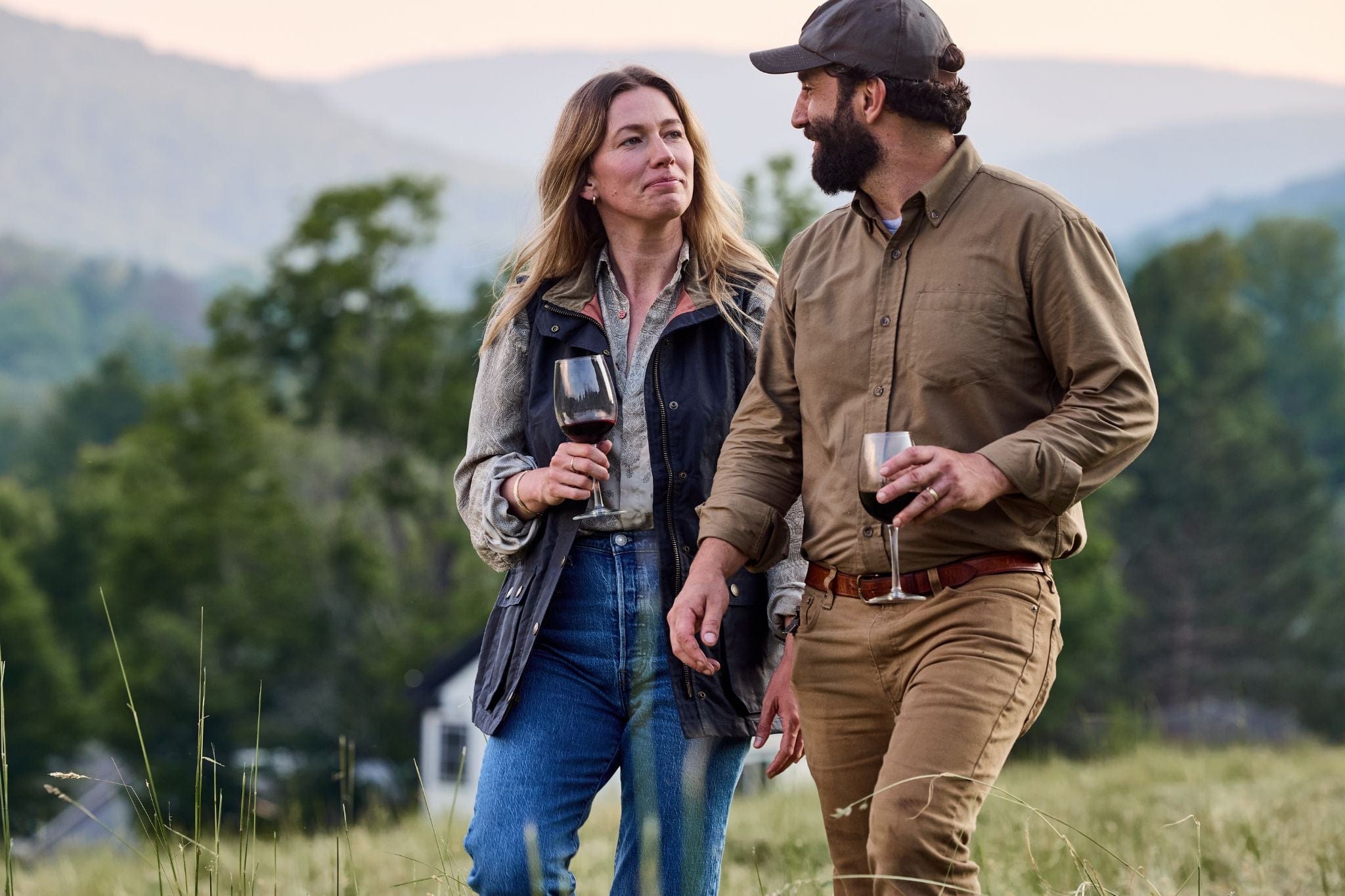 A man and a woman walk together in a grassy field, each holding a drink, with hills and trees in the background.