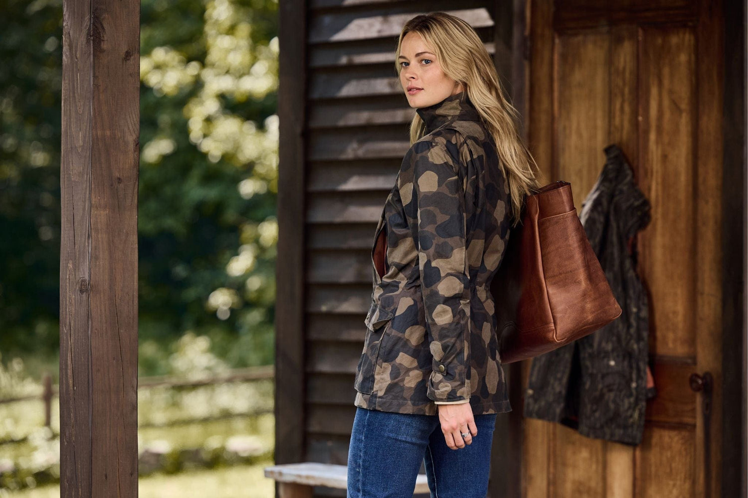 Woman in a camo jacket standing on a porch, carrying a brown bag, with a wooden building and greenery in the background.