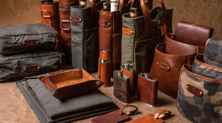 Various leather and canvas bags, flasks, pouches, and trays are neatly arranged on a brown surface.
