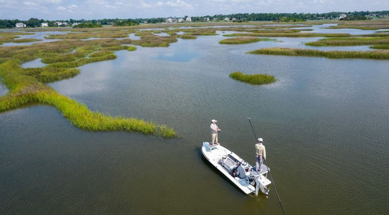 Two people fish on a small boat in a marshy area with scattered patches of grass under a partly cloudy sky.