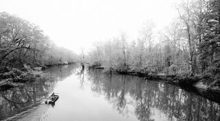 A small boat navigates a calm river surrounded by leafless trees on both banks, reflecting softly on the waters surface.