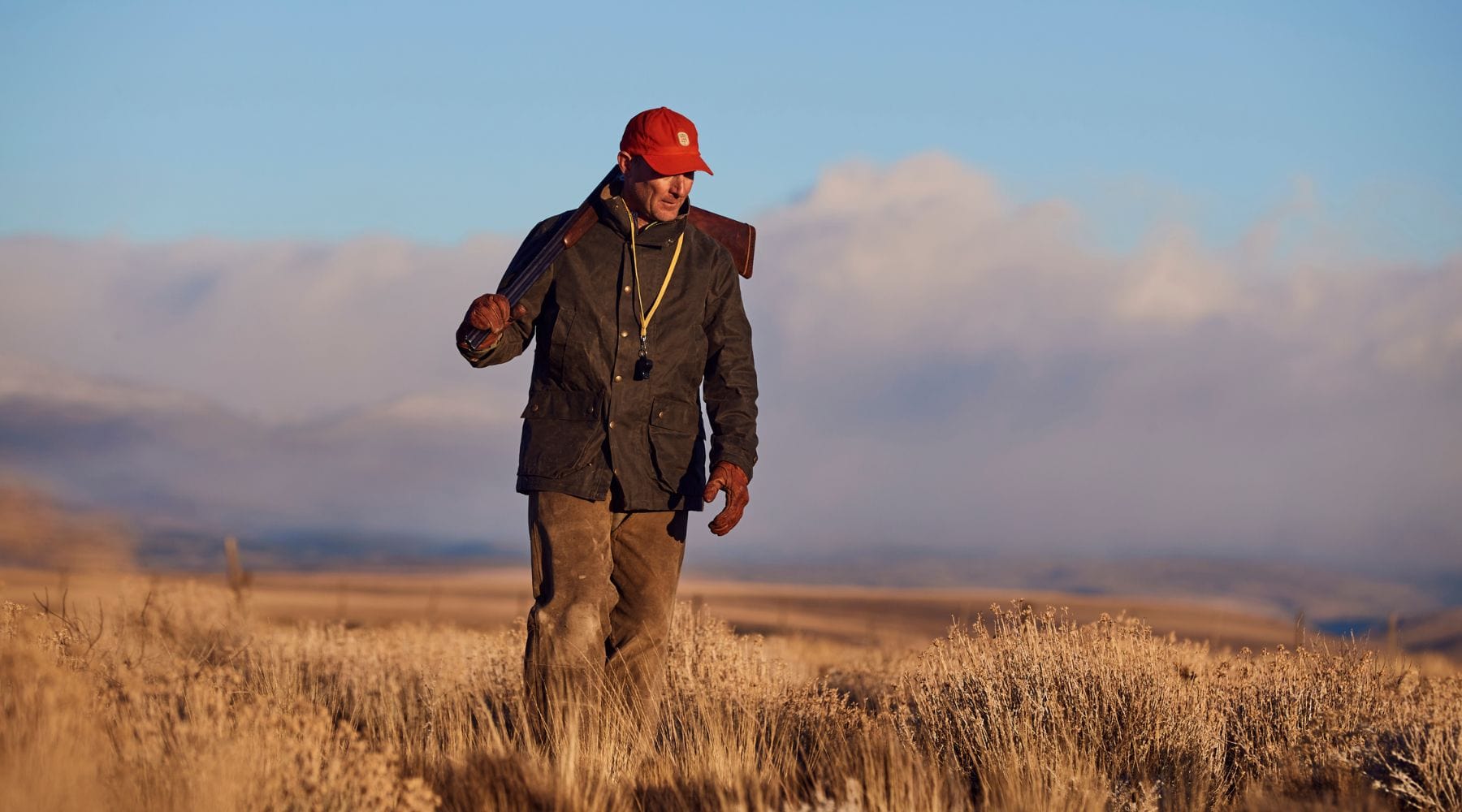 Man in outdoor attire walks through field with a shotgun slung over his shoulder, under a cloudy sky.