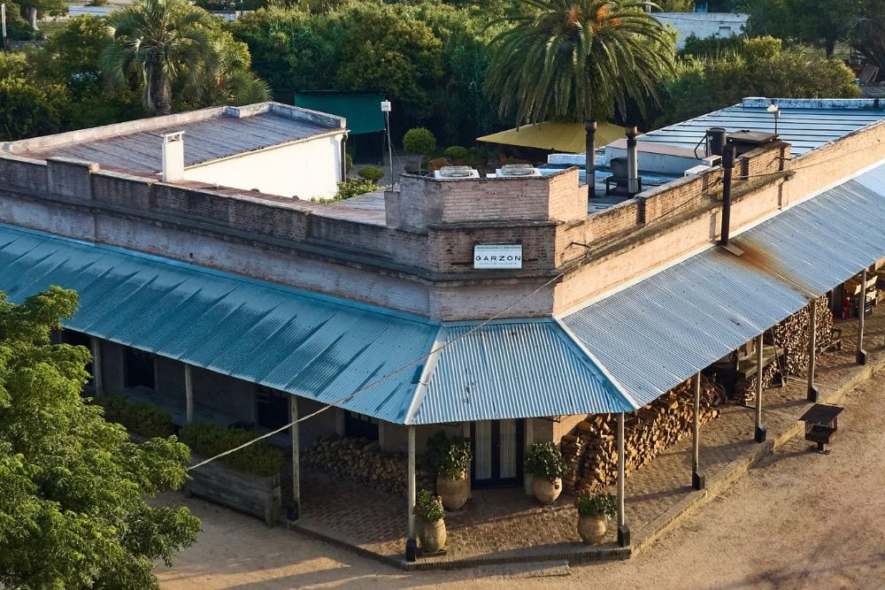 Aerial view of a rustic building with a blue tin roof, surrounded by greenery and a stack of firewood along one side.