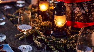 A glass of wine on a wooden table with lit lanterns and greenery in the background during an outdoor setting.
