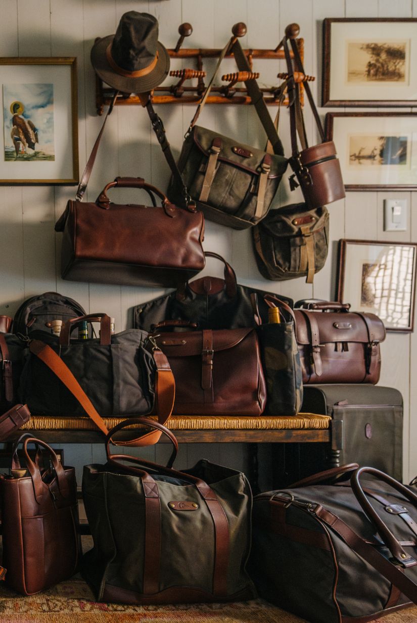 Assorted leather and canvas bags, hats, and framed pictures arranged on a wall, bench, and hooks in a cozy room.