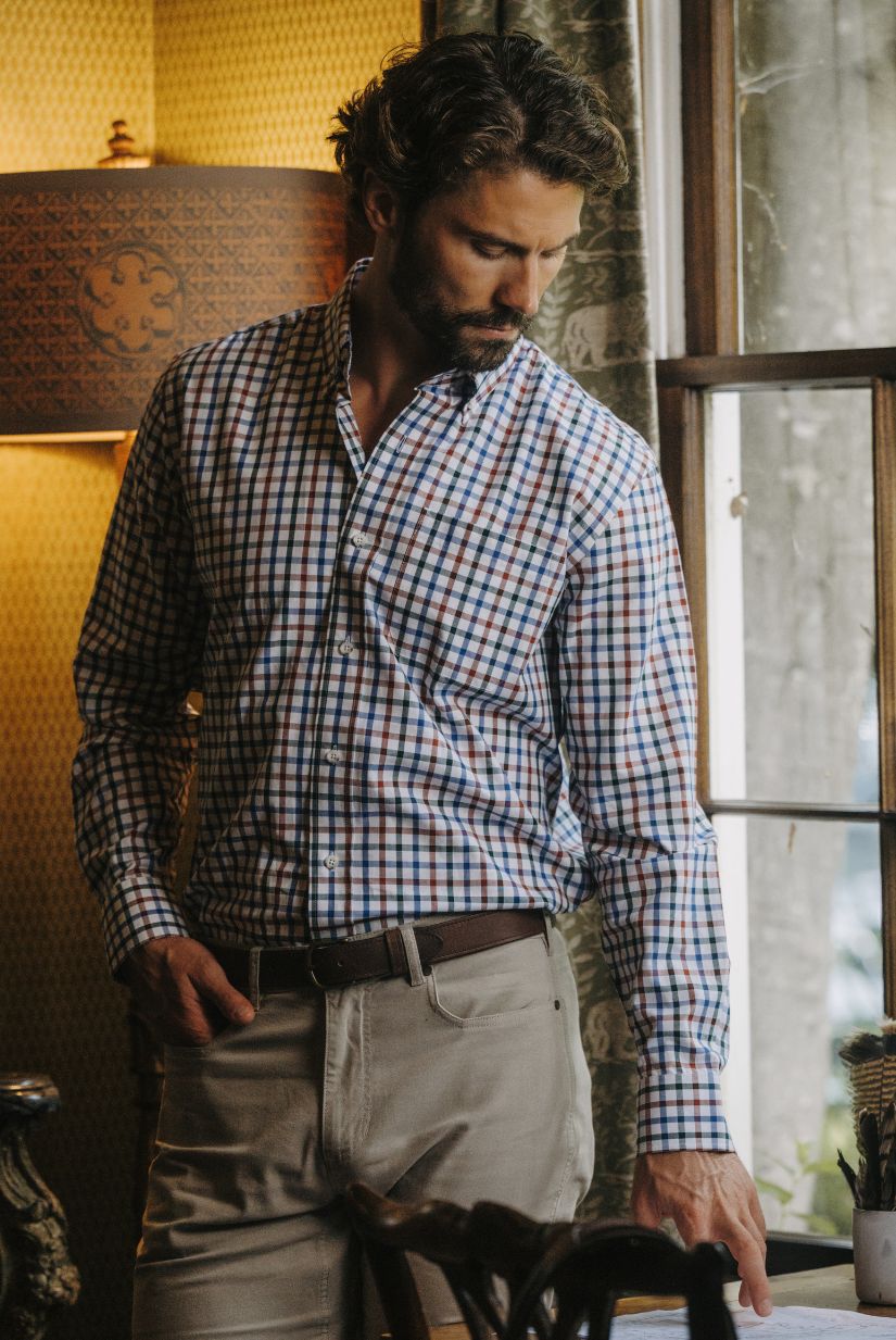 Man in a plaid shirt and khaki pants stands indoors near a window, looking down with one hand on a table.