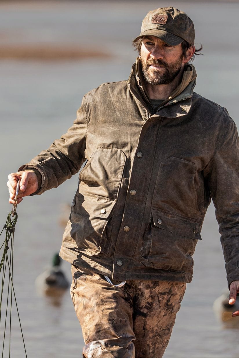 A man in a brown jacket and cap stands outdoors by water, holding rope or cords in one hand.
