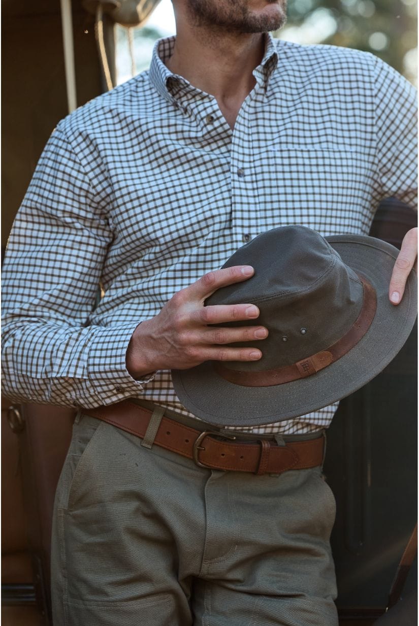 Man in checked shirt and khaki pants holding a hat outdoors.